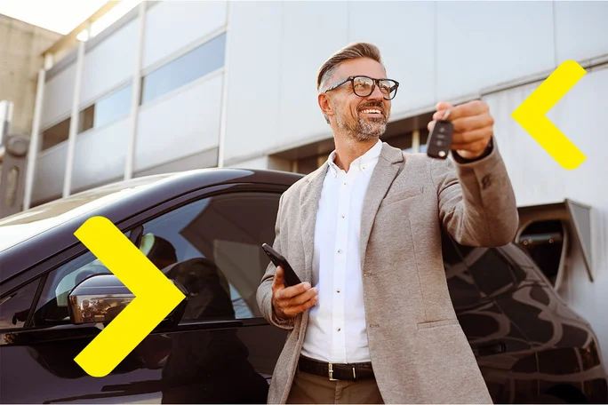 Gospodin drži ključeve automobila u jednoj ruci i pametni telefon u drugoj, stojeći pored crnog automobila