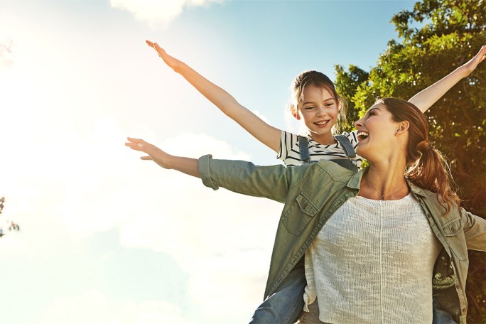 Osoba nosi dijete na ramenima, oboje s ispruženim rukama, uživajući u sunčanom danu na otvorenom
